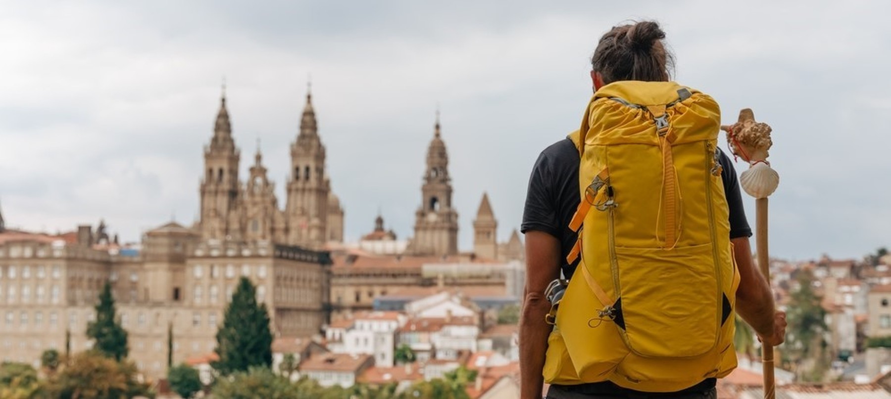 Peregrino con una mochila amarilla y un bastón con la concha de Santiago observando la catedral de Santiago de Compostela