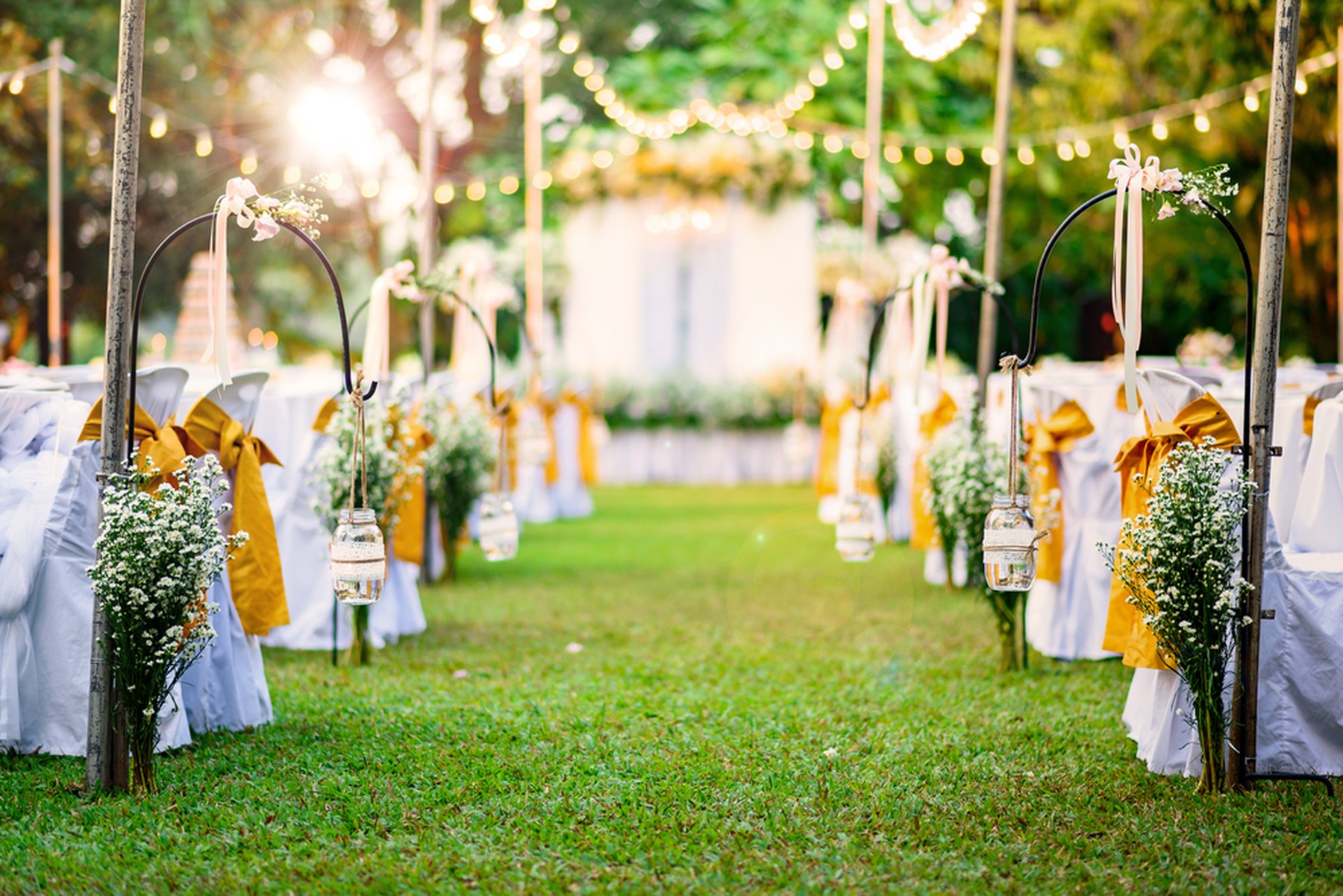 Lugar al aire libre donde se va a celebrar una boda. Se ven guirnaldas de luces, sillas blancas con lazos amarillos, ramos de flores y un altar al fondo.