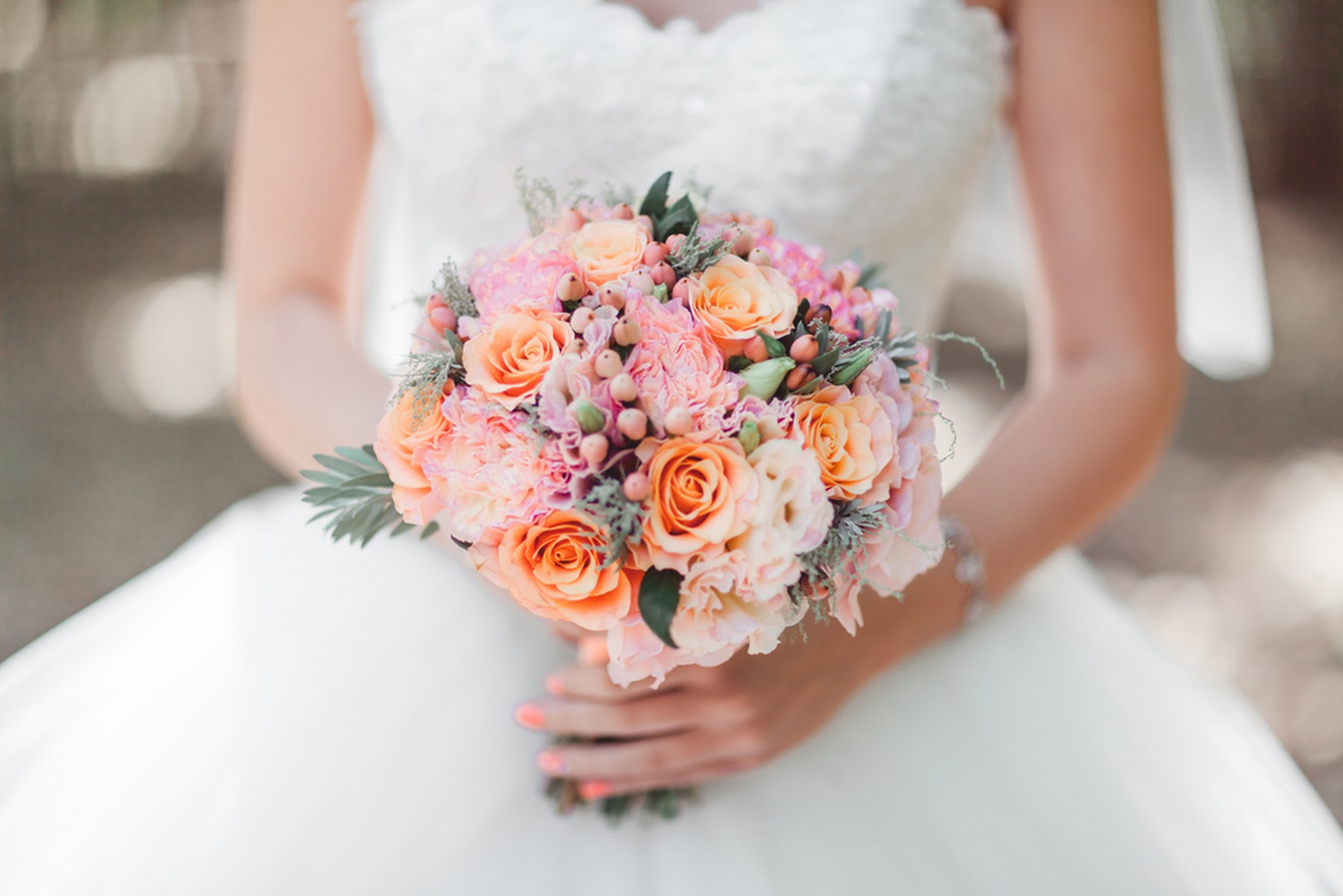 Ramo de flores de novia en tonos rosas y naranjas en primer plano. El ramo lo sujeta la novia, que se ve de fondo su vestido blanco y las uñas pintadas de rosa